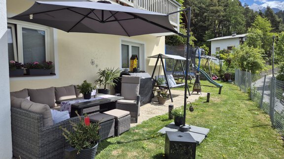 Ein gemütlicher Außenbereich mit Gartenmöbeln und einem Sonnenschirm. Im Hintergrund sind Spielgeräte und ein gepflegter Garten zu sehen. - Alpenlove - Oetz
