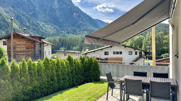Eine gemütliche Terrasse mit einem Esstisch und Stühlen, umgeben von einem gepflegten Rasen und grünen Büschen. Im Hintergrund sind Berge und ein klarer Himmel zu sehen. - ALPsmart - Oetz
