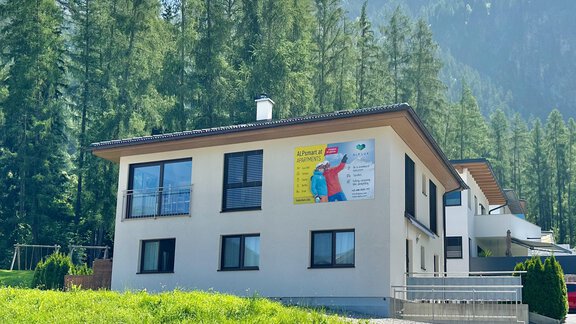 Ein modernes Gebäude umgeben von Bäumen und Bergen. Die Fassade zeigt ein großes Werbebanner. - ALPsmart - Oetz