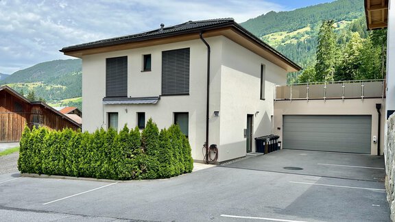 Ein modernes Haus mit einem gepflegten Vorgarten. Im Hintergrund sind grüne Berge und ein blauer Himmel sichtbar. - ALPsmart - Oetz