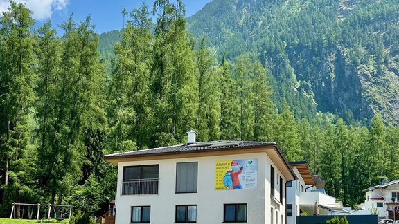 Ein modernes Gebäude umgeben von grünen Bäumen und bergen. Im Hintergrund sind hohe Berge und ein blauer Himmel sichtbar. - ALPsmart - Oetz