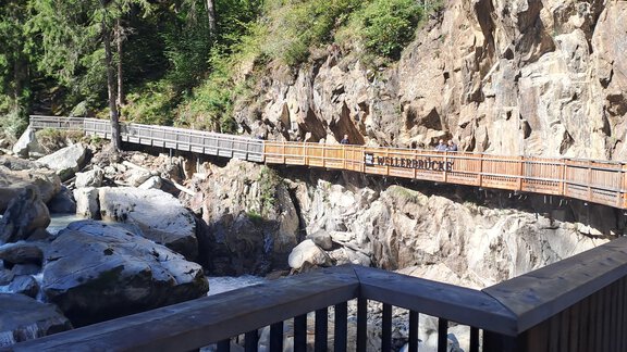 A wooden bridge runs along a rock wall over a river. The surroundings are characterized by trees and natural beauty. - Apart Annabell - Oetz