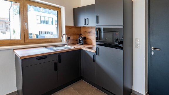 A modern kitchen with black furniture and light wood. It features a sink, a coffee machine, and a microwave. - Appartement Schmied Hof - Oetz