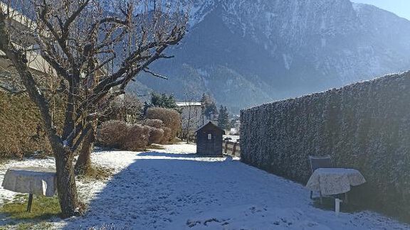 A snowy landscape with a clear blue sky and snow-covered mountains in the background. In the foreground, there is a tree planting and some garden furniture. - Haus Evi - Oetz