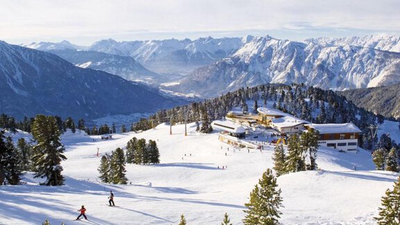 A snowy mountain landscape with skiers on the slope. In the background, majestic mountains and a holiday home are visible. - Haus Evi - Oetz