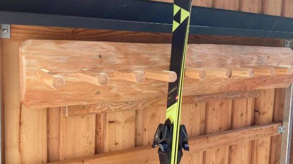 A pair of skis is attached to a wooden rack. In the background, wooden walls and green foliage are visible. - Haus Evi - Oetz