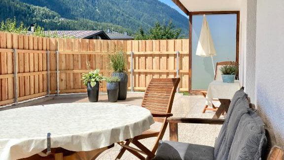 A cozy terrace with a round table and chairs. In the background, mountains and a blue sky can be seen. - Haus Evi - Oetz