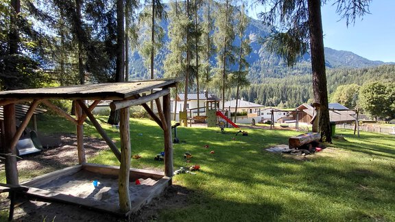 A playground in a green environment with wooden play equipment and a view of the mountains. The sun shines through the trees, creating an inviting atmosphere. - Haus Gstrein - Oetz