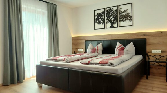 A cozy bedroom with a large bed and decorative cushions. The windows are fitted with a light curtain, and the walls have friendly wood details. - Haus Gstrein - Oetz
