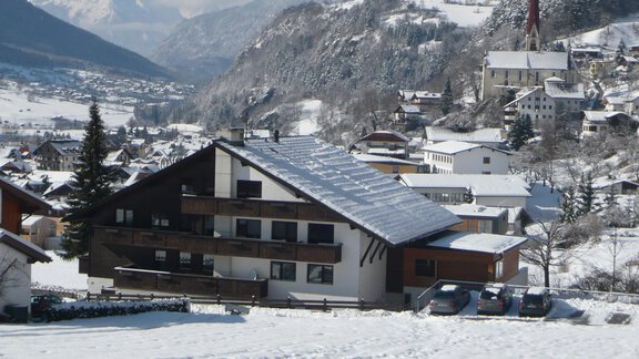 Eine schneebedeckte Landschaft mit einem modernen Wohnhaus und weiteren Gebäuden im Hintergrund. Die umliegenden Berge sind deutlich sichtbar und der Himmel ist klar und blau. - Hotel Marko - Oetz
