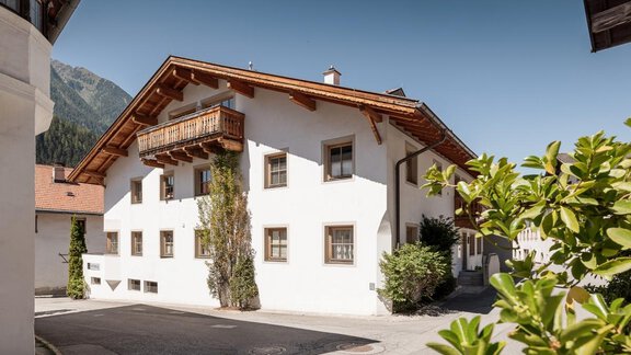 Ein charmantes altes Haus mit einem Holzbalkon in einer ruhigen Straße. Umgeben von grünen Pflanzen und Bergen im Hintergrund. - MYALPS Historical - Oetz