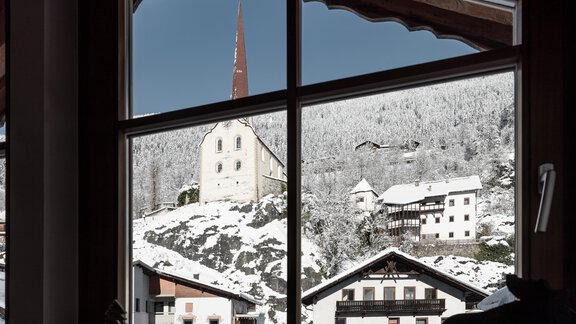 Ein Blick durch ein Fenster auf ein verschneites Dorf mit einer Kirche und mehreren traditionellen Häusern. Die Landschaft ist winterlich und malerisch. - MYALPS Historical - Oetz