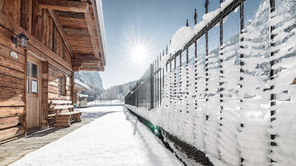 Ein Winterlandschaft mit einem Holzhaus und einer schneebedeckten Umgebung. Die Sonne scheint hell über dem schneebedeckten Zaun. - MYALPS Mühlhof - Oetz