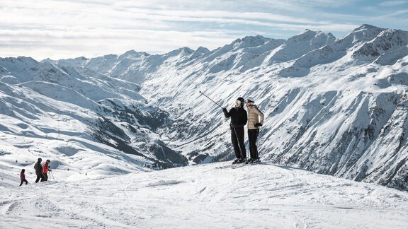 Zwei Skifahrer stehen auf einem schneebedeckten Gipfel und genießen die Aussicht. Im Hintergrund sind beeindruckende Berglandschaften sichtbar. - MYALPS Mühlhof - Oetz