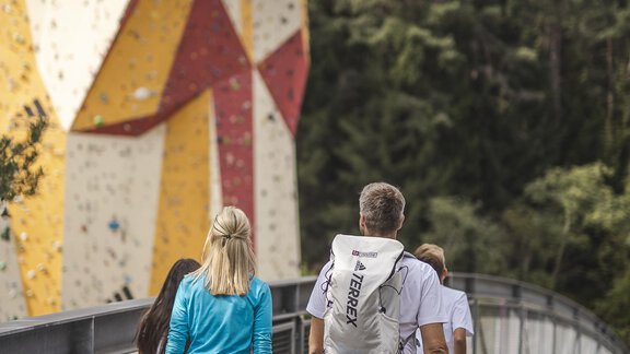 A group of people is walking on a wooden walkway. In the background, there is a colorful climbing wall. - MYALPS Tirol - Oetz