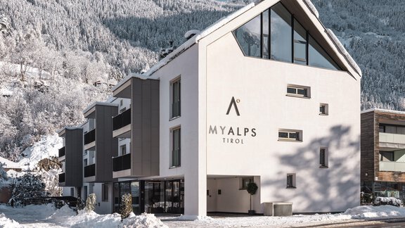 Ein modernes Gebäude mit einer weißen Fassade, umgeben von schneebedeckten Bergen. Die Umgebung ist winterlich und idyllisch. - MYALPS Tirol - Oetz