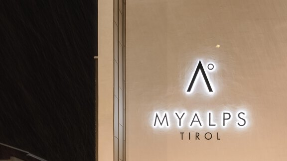 Ein modernes Gebäude mit einem beleuchteten Schild für "MYALPS TIROL". Die Umgebung ist schneebedeckt und wirkt winterlich. - MYALPS Tirol - Oetz