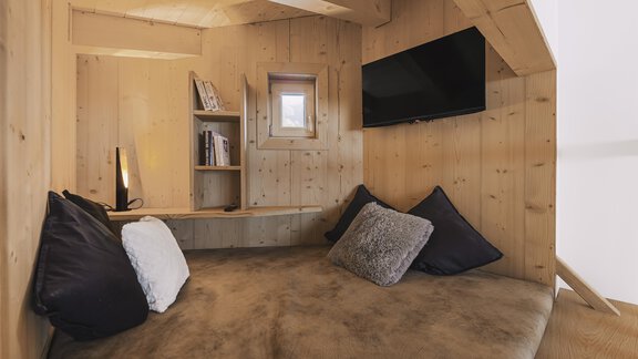 Ein gemütlicher Raum aus Holz mit einem großen Sitzbereich. An der Wand hängt ein Fernseher und es gibt ein Regal mit Büchern. - See das Ferienhaus - Oetz