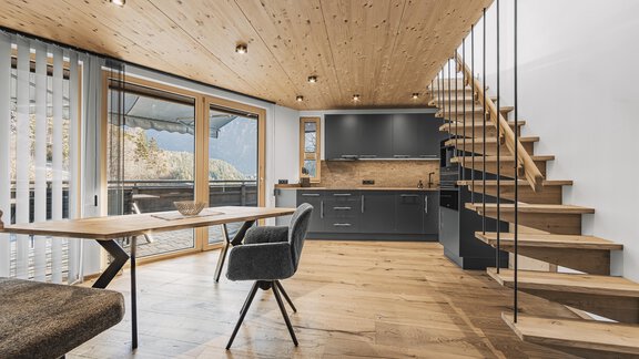 Eine modern gestaltete Wohnküche mit einer Holztreppe und einem großen Fenster, das viel Licht hereinlässt. Der Raum ist mit einem Esstisch und stilvollen Möbeln eingerichtet. - See das Ferienhaus - Oetz