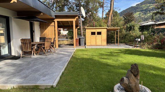 A beautiful garden with a terrace, a dining table, and chairs. In the background, there is a small garden house, and the landscape is surrounded by trees and mountains. - Residenz Bergzauber - Ötztal Bahnhof