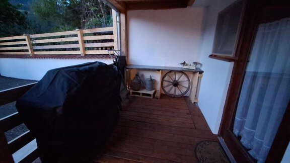 A terrace with a wooden floor, a grill, and a table. In the background, a wagon wheel and an opaque wall are visible. - Appartement Harmony - Sautens
