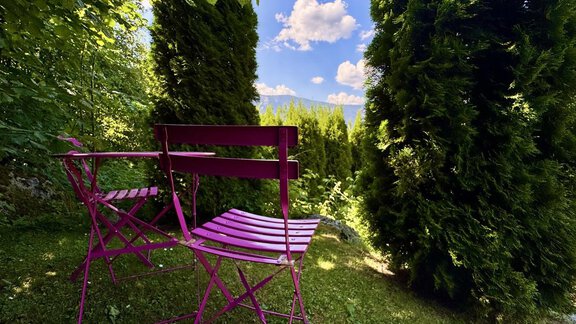 Zwei pinke Stühle stehen im Garten, umgeben von hohen grünen Bäumen. Der Himmel ist blau mit einigen Wolken und die Landschaft wirkt einladend und ruhig. - bei Herrmann´s - Sautens