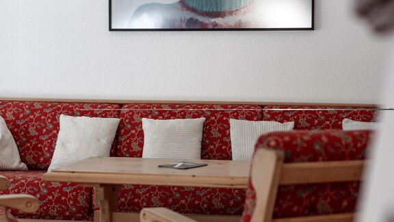 Ein gemütlicher Wohnraum mit einem Holztisch und roten Polstermöbeln. An der Wand hängt ein Bild einer Person mit Skibrille vor einer Schneelandschaft. - Chalet Pirchhof by MYALPS - Sautens