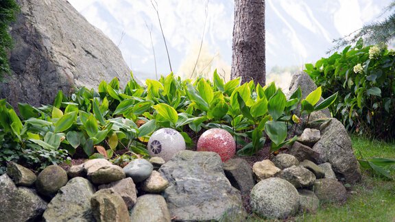 Ein kleiner Gartenbereich mit grünen Pflanzen und bunten Kugeln. Felsen umrahmen das Pflanzenbett und im Hintergrund sind Berge zu sehen. - Ferienwohnungen Inka - Sautens