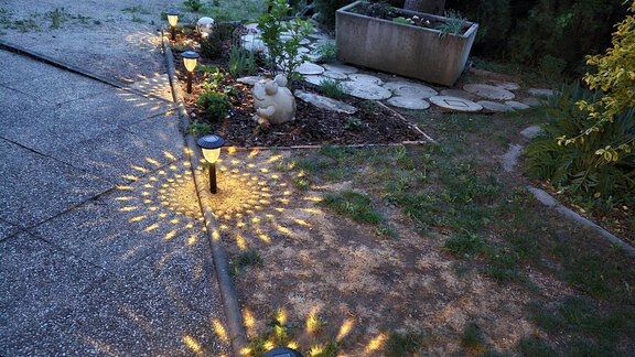 Ein schöner Gartenweg mit leuchtenden Gartenleuchten, die ein Muster auf den Boden werfen. Im Hintergrund sind ein Auto und einige Pflanzen zu sehen. - Ferienwohnungen Inka - Sautens