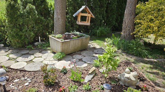 Ein gepflegter Garten mit Steinstufen und bunten Blumen. Im Hintergrund steht ein Vogelhaus in den Bäumen. - Ferienwohnungen Inka - Sautens