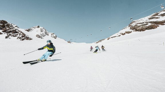 A group of skiers is skiing down a snow-covered slope. The sky is clear and blue, and the mountains in the background are visible. - A CASA "Aventurin" - Sölden