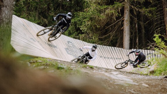 Three mountain bikers ride on a winding trail in the forest. The surroundings are green and natural, with trees in the background. - A CASA "Aventurin" - Sölden