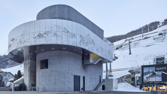 A modern building with a unique architectural shape stands in the midst of a snowy landscape. In the background, ski slopes and a ski lift are visible. - A CASA "Aventurin" - Sölden