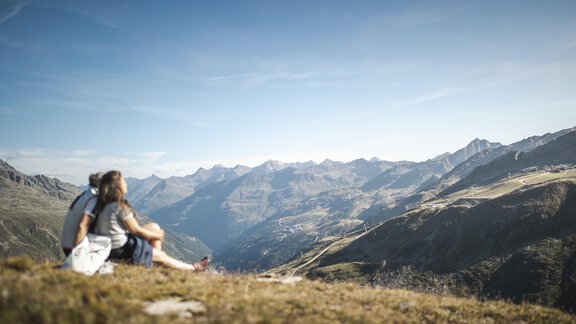 A couple is sitting on a hill, enjoying the view of the mountains. The sky is clear and the landscape is green and hilly. - A CASA "Aventurin" - Sölden