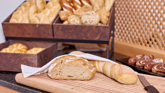 A selection of fresh bread and rolls in wooden boxes. On a cutting board, there is a sliced loaf and an elongated roll. - Alp Resort Tiroler Adler - Sölden