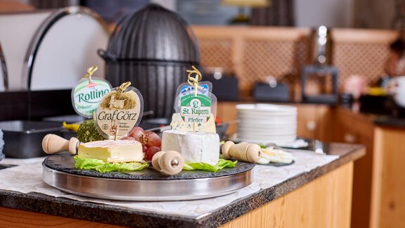 A beautifully arranged cheese cart with various cheese types on a table. In the background, dishes and a warm atmosphere can be seen. - Alp Resort Tiroler Adler - Sölden