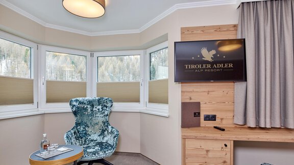 A modern room with large windows and a comfortable armchair. There is a television on the wall as well as a wooden desk. - Alp Resort Tiroler Adler - Sölden
