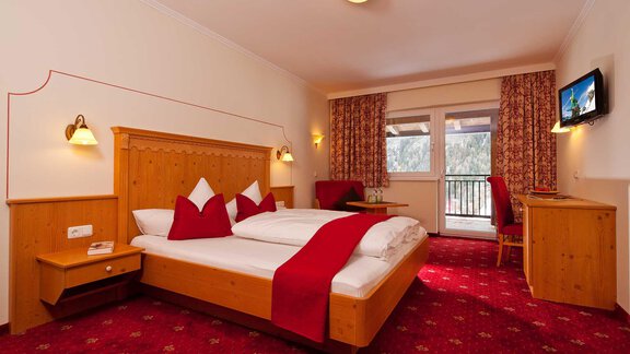 A cozy hotel room with a double bed and red accents. Wooden paneling hangs on the walls and there are large windows with a view outside. - Alp Resort Tiroler Adler - Sölden