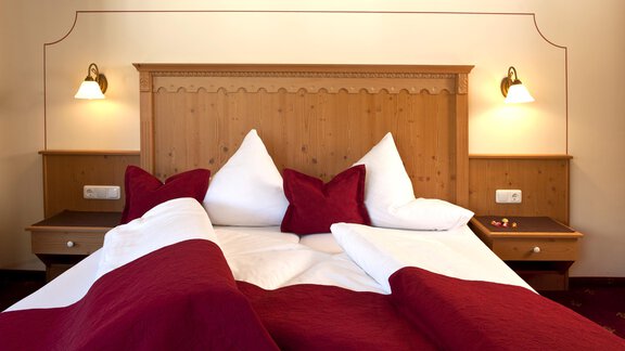 A cozy bedroom with a wooden bed frame and red pillows. The walls are light, and there is soft lighting from wall lamps. - Alp Resort Tiroler Adler - Sölden