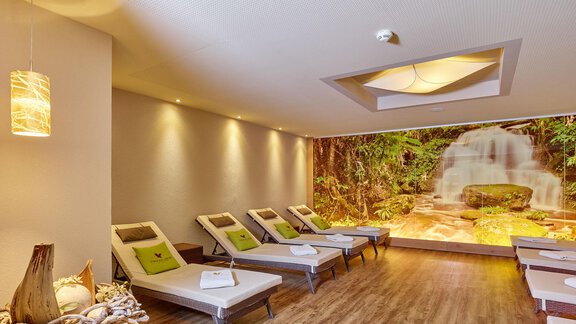 A relaxing wellness area with loungers and a wall featuring a waterfall motif. Soft light and natural elements create a calming atmosphere. - Alp Resort Tiroler Adler - Sölden