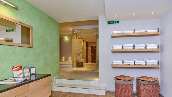 A modern reception area with wooden flooring and a green accent. Shelves with fresh towels and baskets give the space an inviting atmosphere. - Alp Resort Tiroler Adler - Sölden
