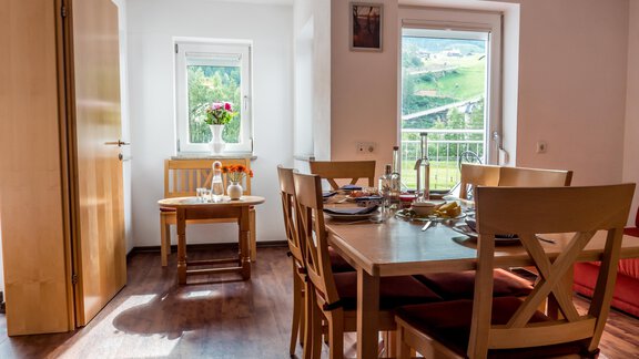 Ein helles und einladendes Esszimmer mit einem Tisch und Stühlen. Durch das Fenster sieht man eine grüne Landschaft und blühende Pflanzen. - Alpenapart Saphir - Sölden