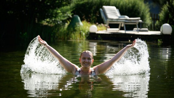 Eine Frau schwimmt lächelnd im klaren Wasser und spritzt mit den Armen. Im Hintergrund sind Liegestühle und grüne Pflanzen zu sehen. - Alpengasthof Grüner - Sölden