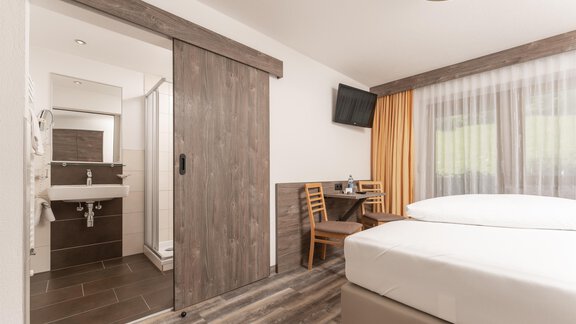 A modern hotel room with a double bed and a small table. In the background, there is a bathroom area and large windows with curtains. - Alpenliebe - Sölden