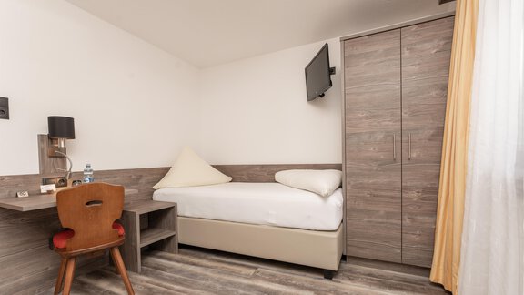 A simple room with a single bed, a desk, and a television. The design is modern and bright, with wood paneling and a large window. - Alpenliebe - Sölden