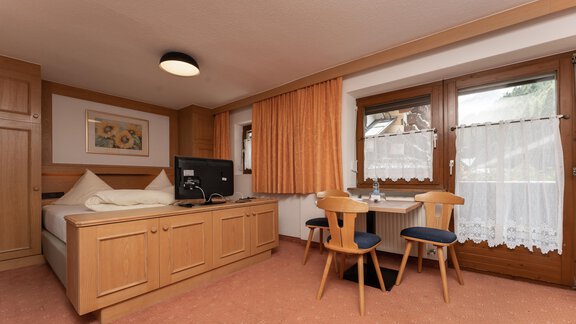 A cozy room with wooden furniture, a double bed, and a table with chairs. Large windows provide plenty of daylight. - Alpenliebe - Sölden
