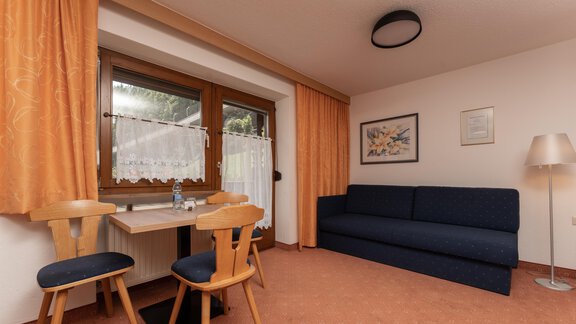 A cozy living space with a blue sofa and a small dining table. Large windows with a view outside and orange curtains give the room a friendly atmosphere. - Alpenliebe - Sölden