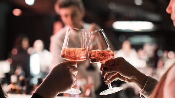 Two glasses with pink liquid clink together. In the background, there are blurred silhouettes of people. - AlpenParks The Secret Sölden - Sölden