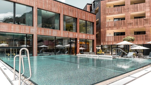 An elegant outdoor pool surrounded by modern buildings.  
Umbrellas and loungers invite you to relax. - AlpenParks The Secret Sölden - Sölden