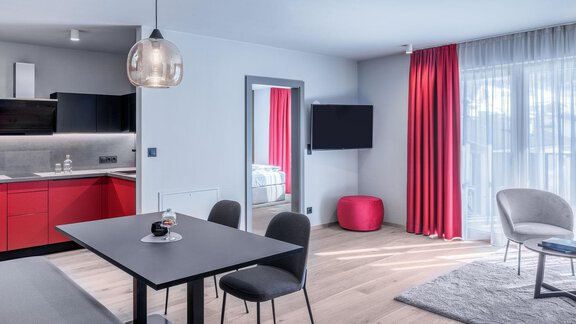 The image shows a modern room with a kitchenette and a dining table. The decor is stylish with red curtains and a cozy seating area. - AlpenParks The Secret Sölden - Sölden